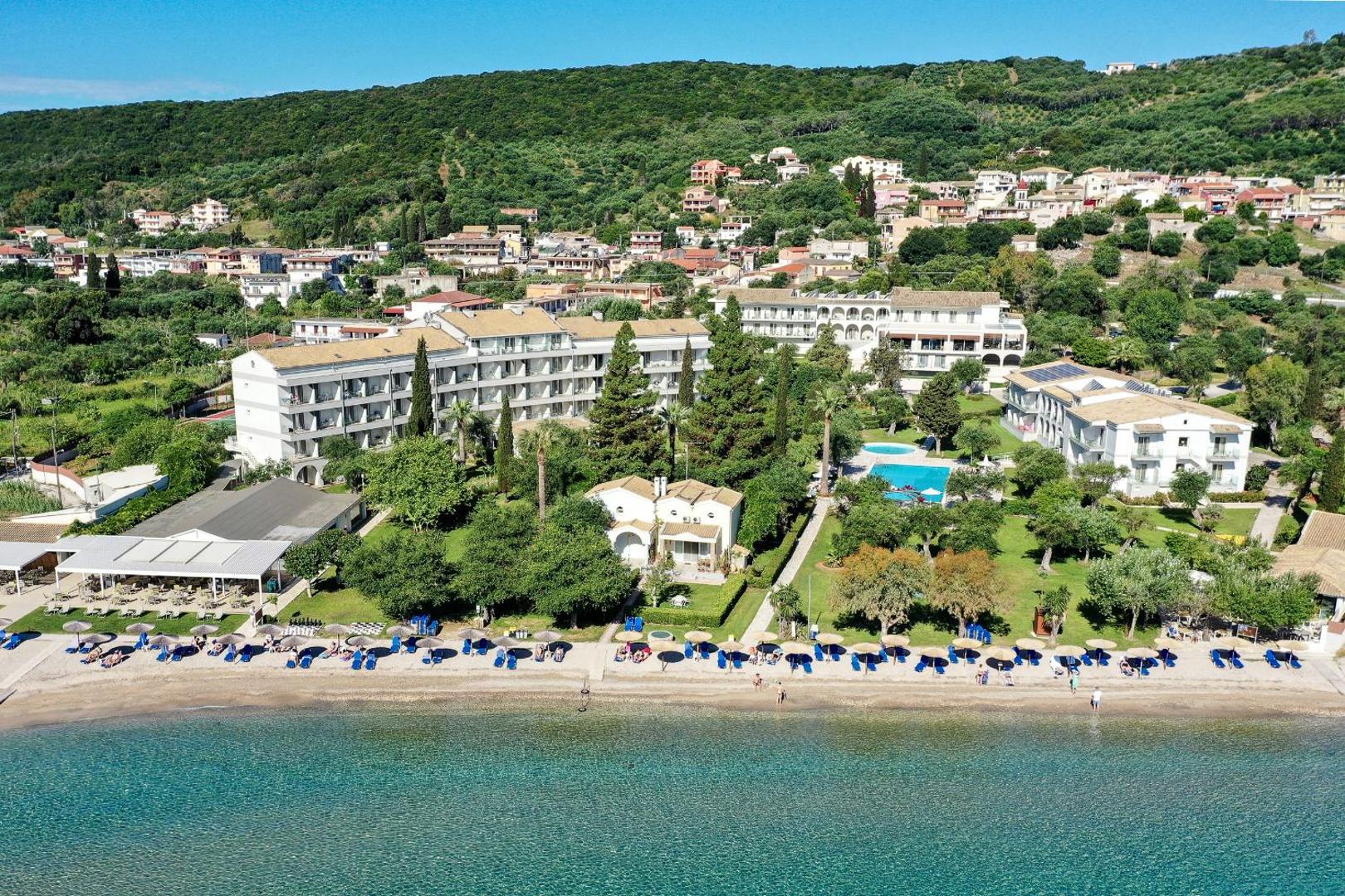 Delfinia Hotel skats no putna lidojuma Korfu salā — viesnīcas ēkas, zaļš dārzs ar baseinu un privātā pludmale pie dzidra ūdens