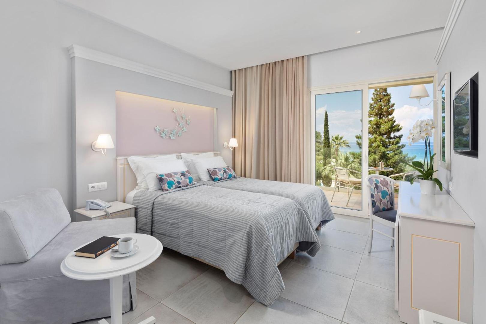 Delfinia Hotel numura interjers ar divām gultām un balkonu, no kura paveras skats uz jūru un priedēm Korfu