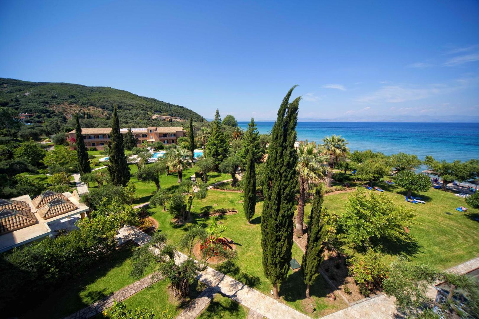 Delfinia Hotel plašā dārza un piekrastes panorāma Korfu salā — skats no augšas uz zaļo teritoriju, olīvkokiem un zilo Jonijas jūru