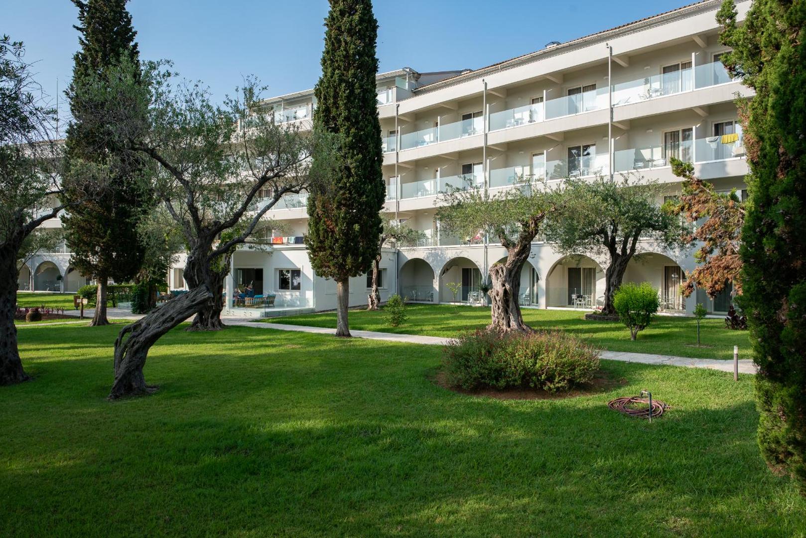Delfinia Hotel numuru korpuss ar balkoniem, ko ieskauj zaļš zāliens un veci olīvkoki dārzā