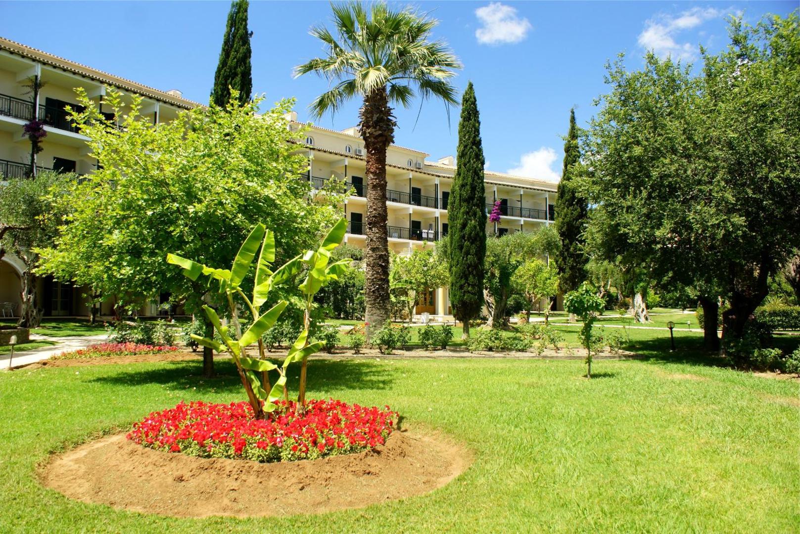 Delfinia Hotel dārza ainava ar palmām, cipresēm un ziedošām puķu dobēm pie viesnīcas ēkas saulainā dienā
