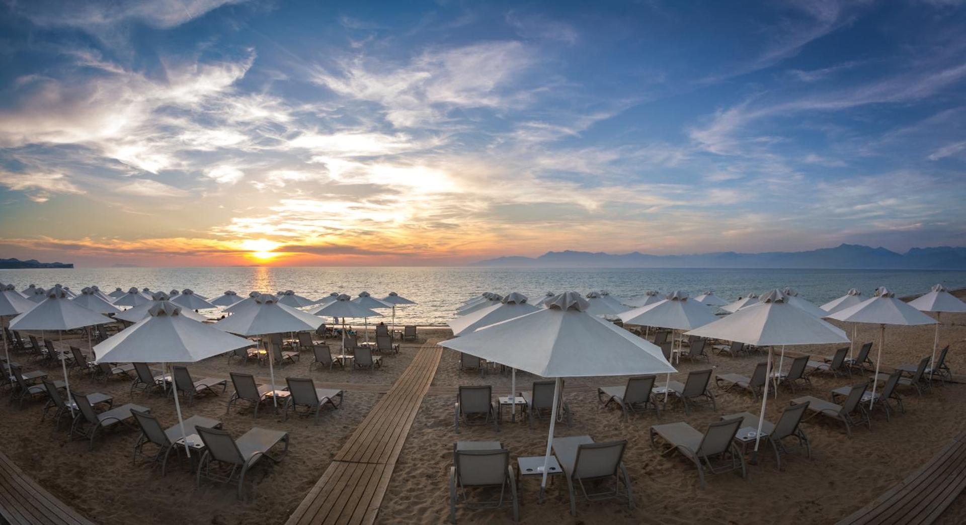 Acharavi Beach viesnīcas pludmales zona ar daudziem baltiem saulessargiem un sauļošanās krēsliem saulrieta laikā Korfu