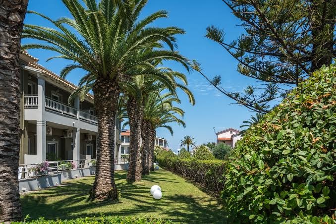 Acharavi Beach viesnīcas modernā ēka ar balkoniem, ko ieskauj augstas palmas un rūpīgi kopts zālājs saulainā dienā.