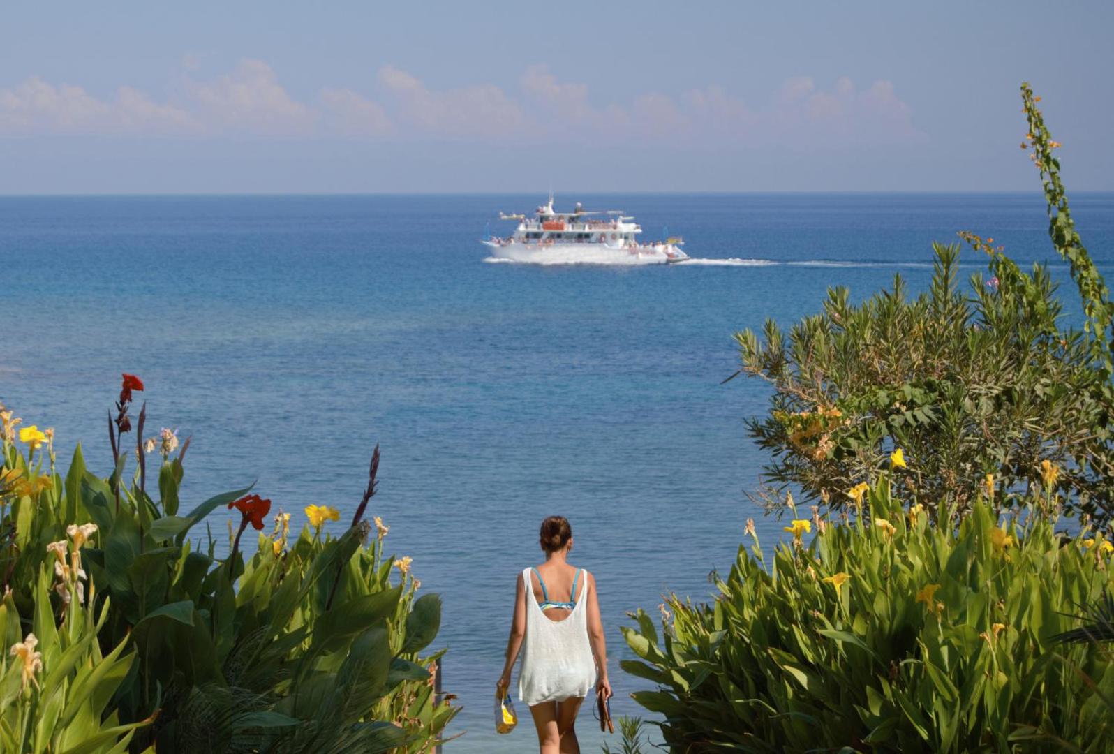 Skats uz Vidusjūru no Crystal Springs Beach viesnīcas dārza — tirkīzzils ūdens, garām braucošs kuģītis un ziedoši augi piekrastē