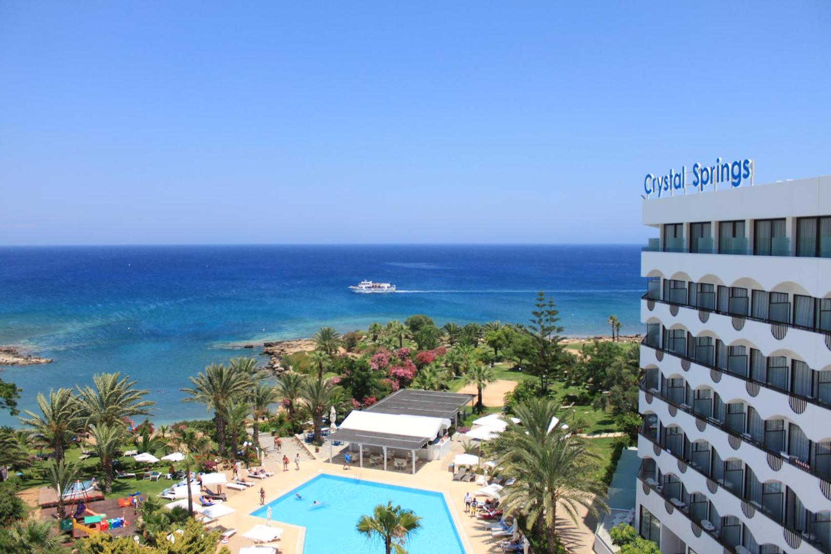 Crystal Springs Beach Hotel ēkas fasāde un teritorija Protarasā — skats uz baseinu, dārzu un zilo Vidusjūru ar kuģi tālumā.