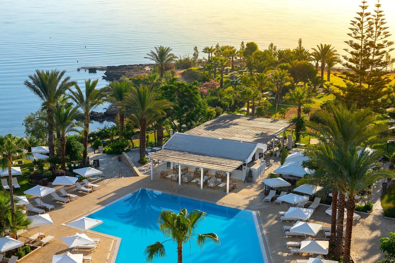 Crystal Springs Beach Hotel teritorijas skats no augšas Protarasā — āra baseins, bārs ar terasi, palmas un Vidusjūra saulrietā.