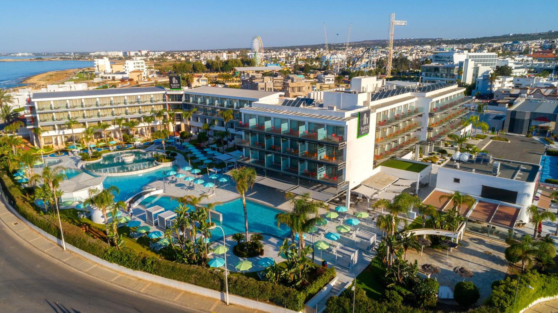 Faros Hotel Ayia Napa viesnīcas ēka un baseinu teritorija uz pilsētas un panorāmas rata fona Kiprā — saulains dienas skats