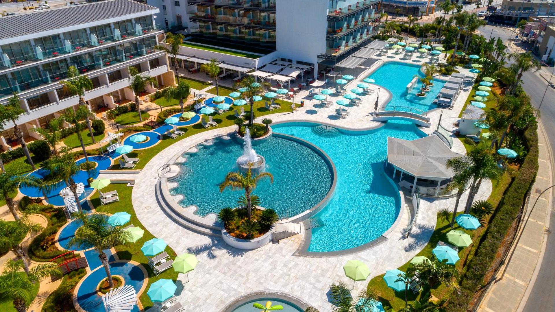 Faros Hotel Ayia Napa plašā baseinu zona ar strūklaku, saulessargiem un palmu dārzu Kiprā — skats no augšas uz atpūtas zonu