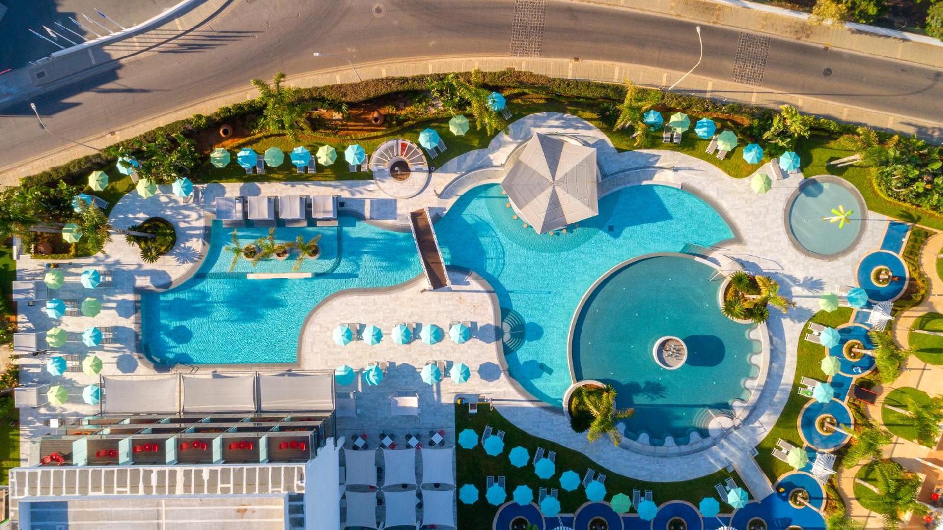 Faros Hotel baseinu kompleksa aerofoto skats no augšas — redzami dažādu formu baseini, tiltiņš, bārs un saulessargu rindas