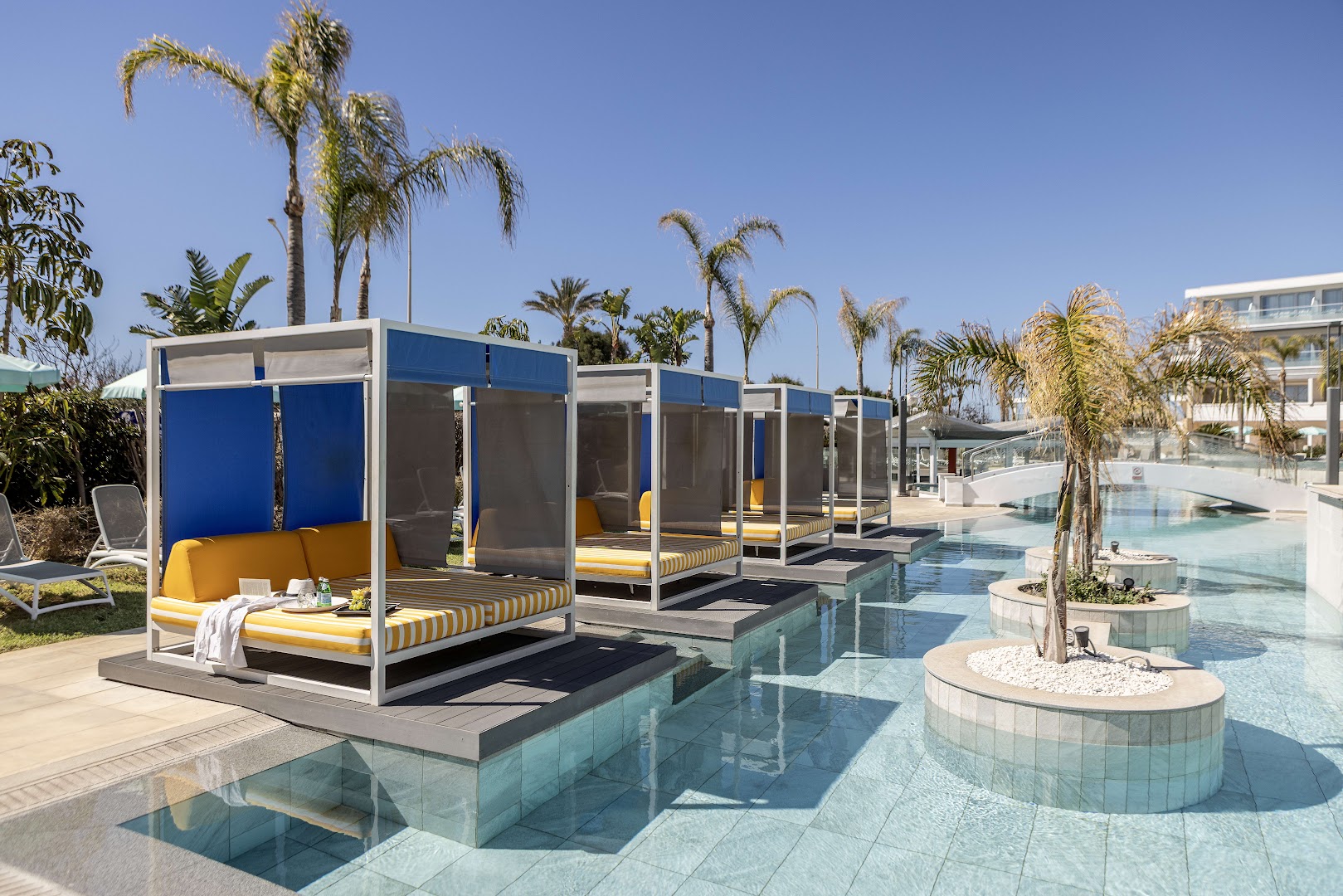 Faros Hotel modernās atpūtas nojumes (cabanas) pie baseina ar dzelteniem matračiem un palmām fonā Ajanapā