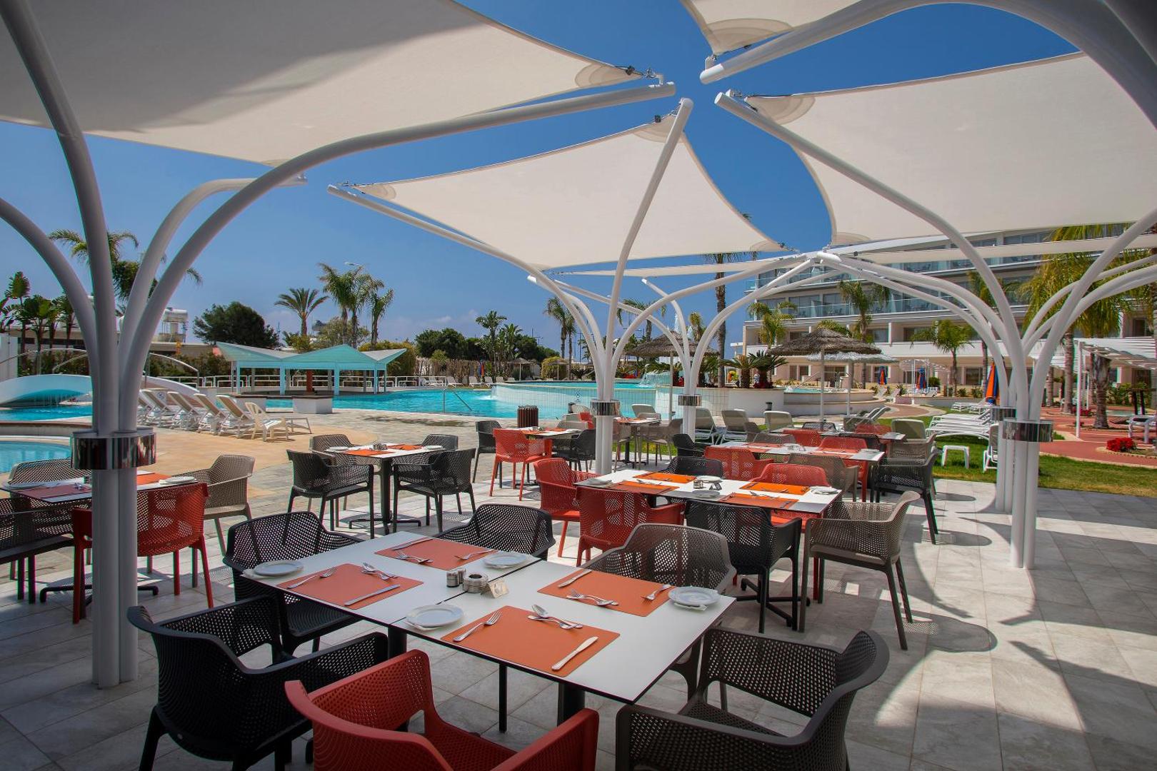 Faros Hotel āra terases restorāns ar modernām saules burām Ajanapā, Kiprā — skats uz baseinu un viesnīcas dārzu saulainā dienā