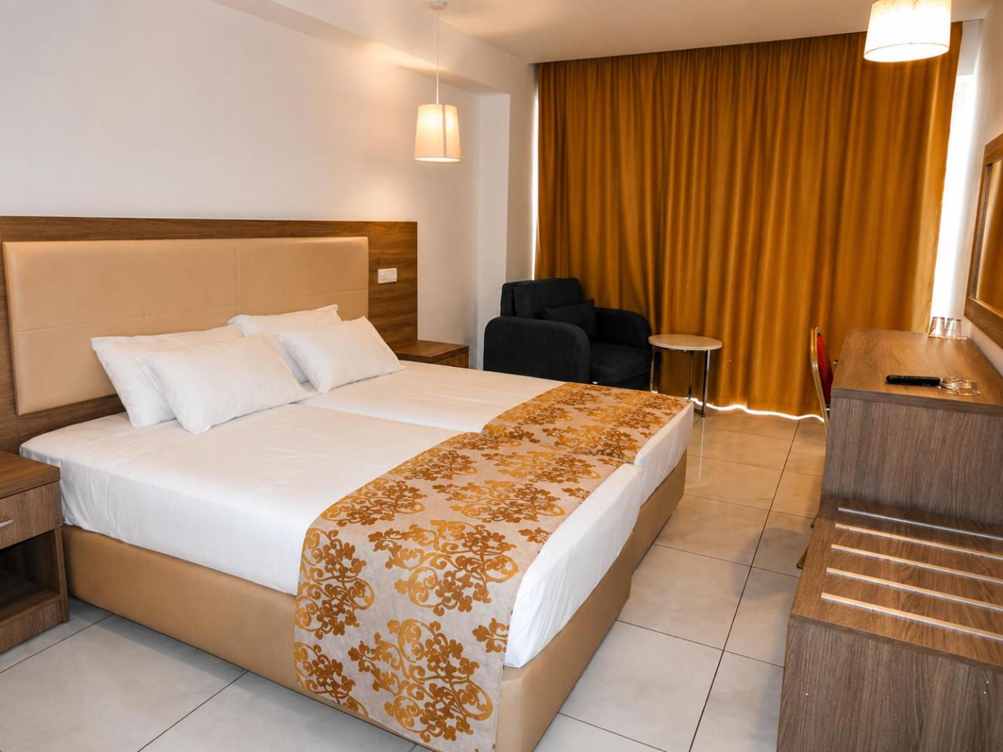 Viesnīcas Corfu Hotel numura interjers ar divām gultām, tumši zilu atpūtas krēslu un mēbelēm koka apdarē