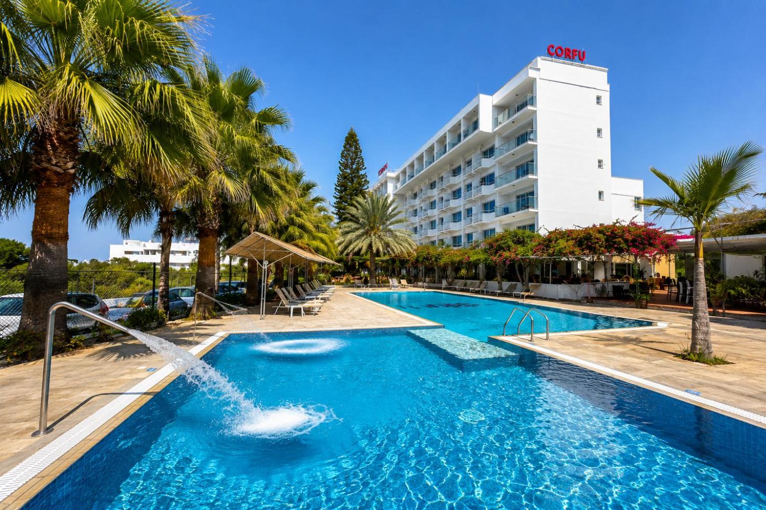 Corfu Hotel Ajanapā, Kiprā — galvenā ēka un āra baseins ar strūklaku, ko ieskauj palmas un atpūtas krēsli saulainā dienā