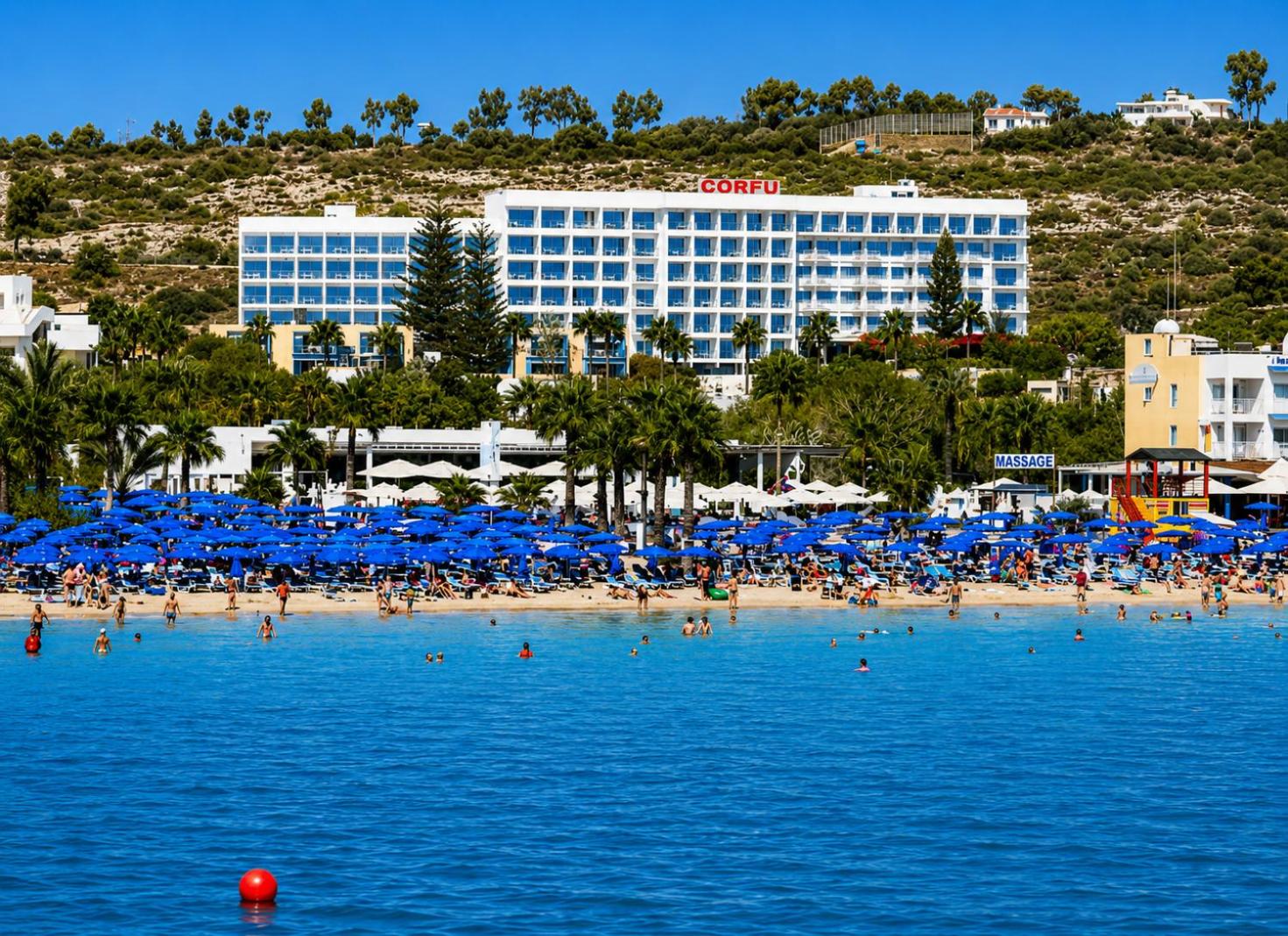 Corfu Hotel skats no jūras puses Aijanapā, Kiprā — balta daudzstāvu ēka piekrastē ar ziliem saulessargiem un atpūtniekiem pludmalē