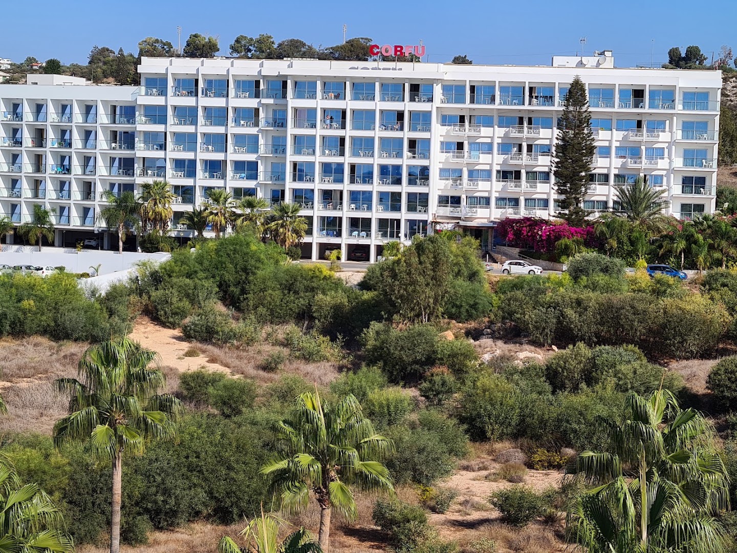 Corfu Hotel fasāde ar stiklotiem balkoniem un apkārtējo zaļo zonu Aijanapā, Kiprā — skats no dārza puses