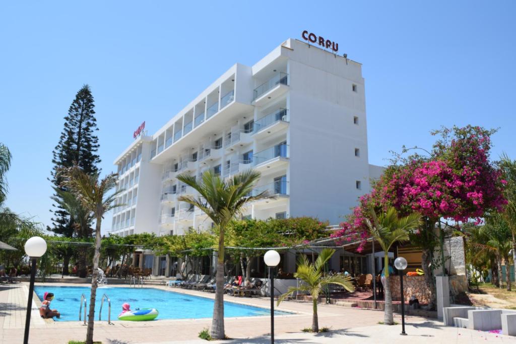 Corfu Hotel baseina zona ar bērnu peldriņķi, sauļošanās krēsliem un ziedošiem krūmiem pie viesnīcas galvenās ēkas Aijanapā