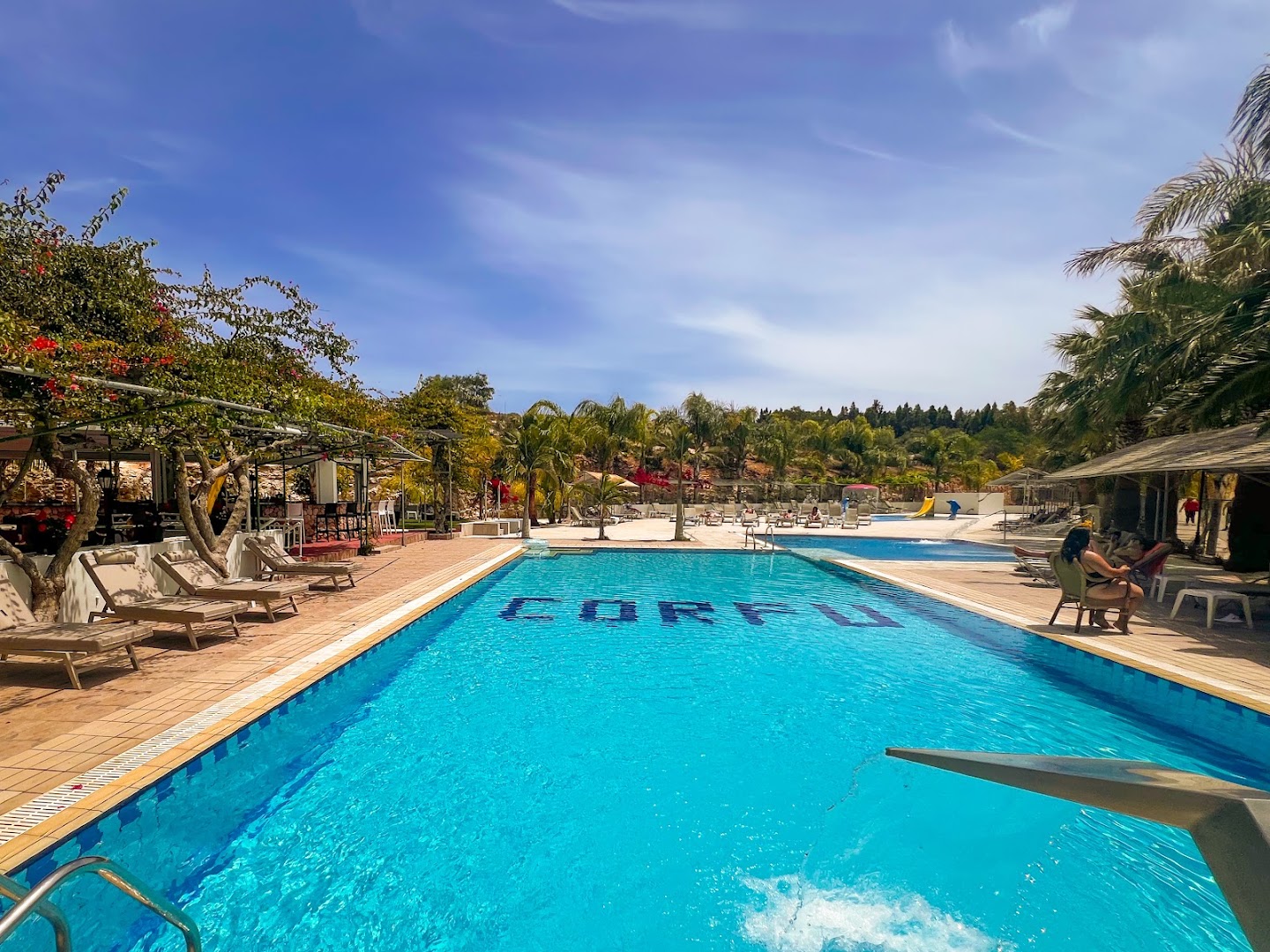 Corfu Hotel peldbaseins ar skatu uz dārzu un atpūtas krēsliem Ajanapā, Kiprā — saulains pagalms ar ziedošiem krūmiem