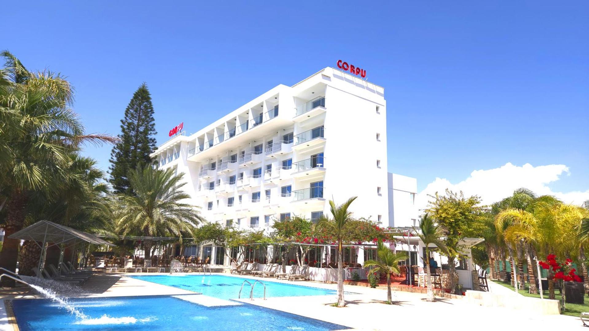 Corfu Hotel āra baseins ar zilu ūdeni, ko ieskauj palmas un viesnīcas baltā fasāde ar balkoniem Aijanapā