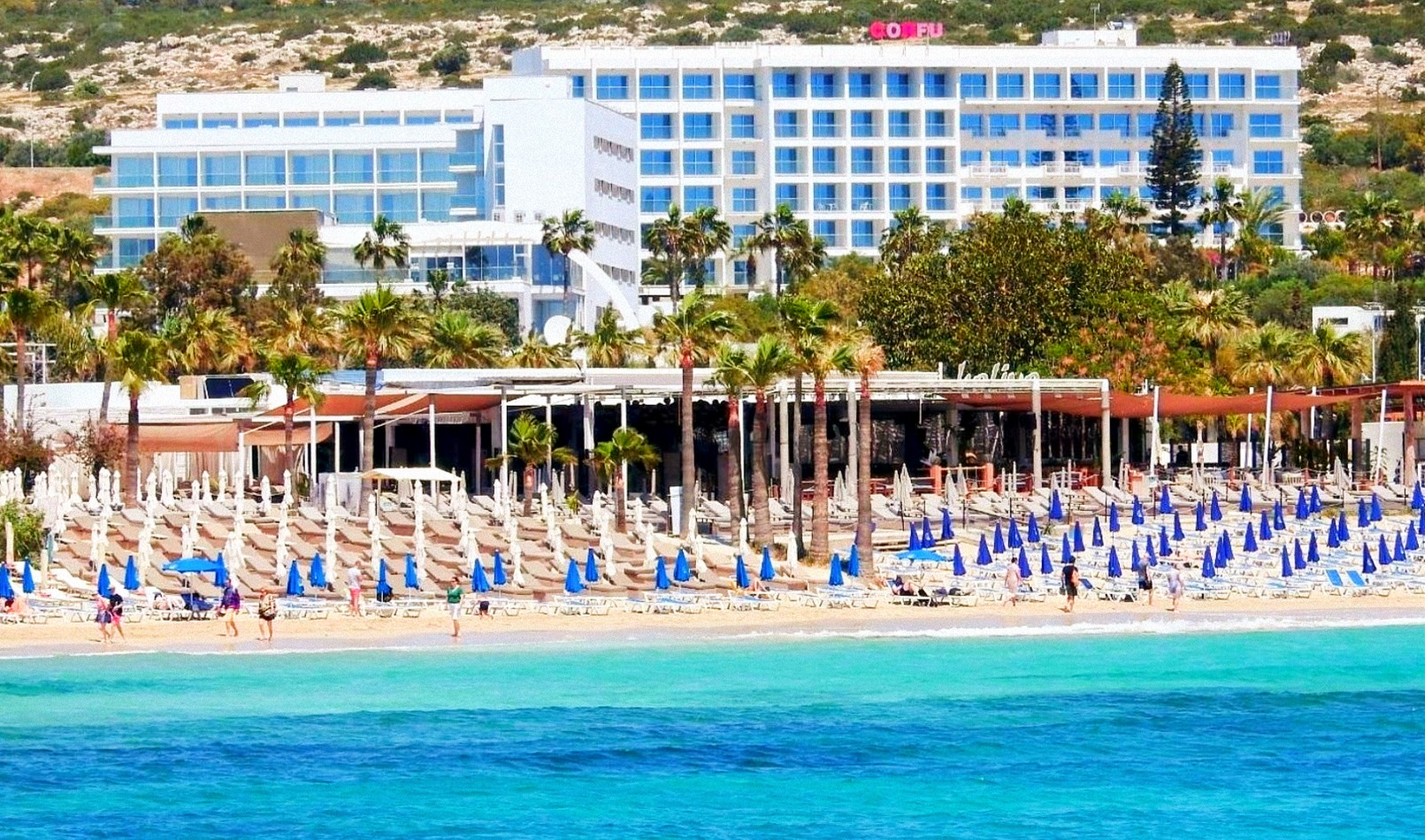 Corfu Hotel Aijanapā skats uz pludmales zonu ar daudziem sauļošanās krēsliem, saulessargiem un palmu rindu pie viesnīcas ēkas