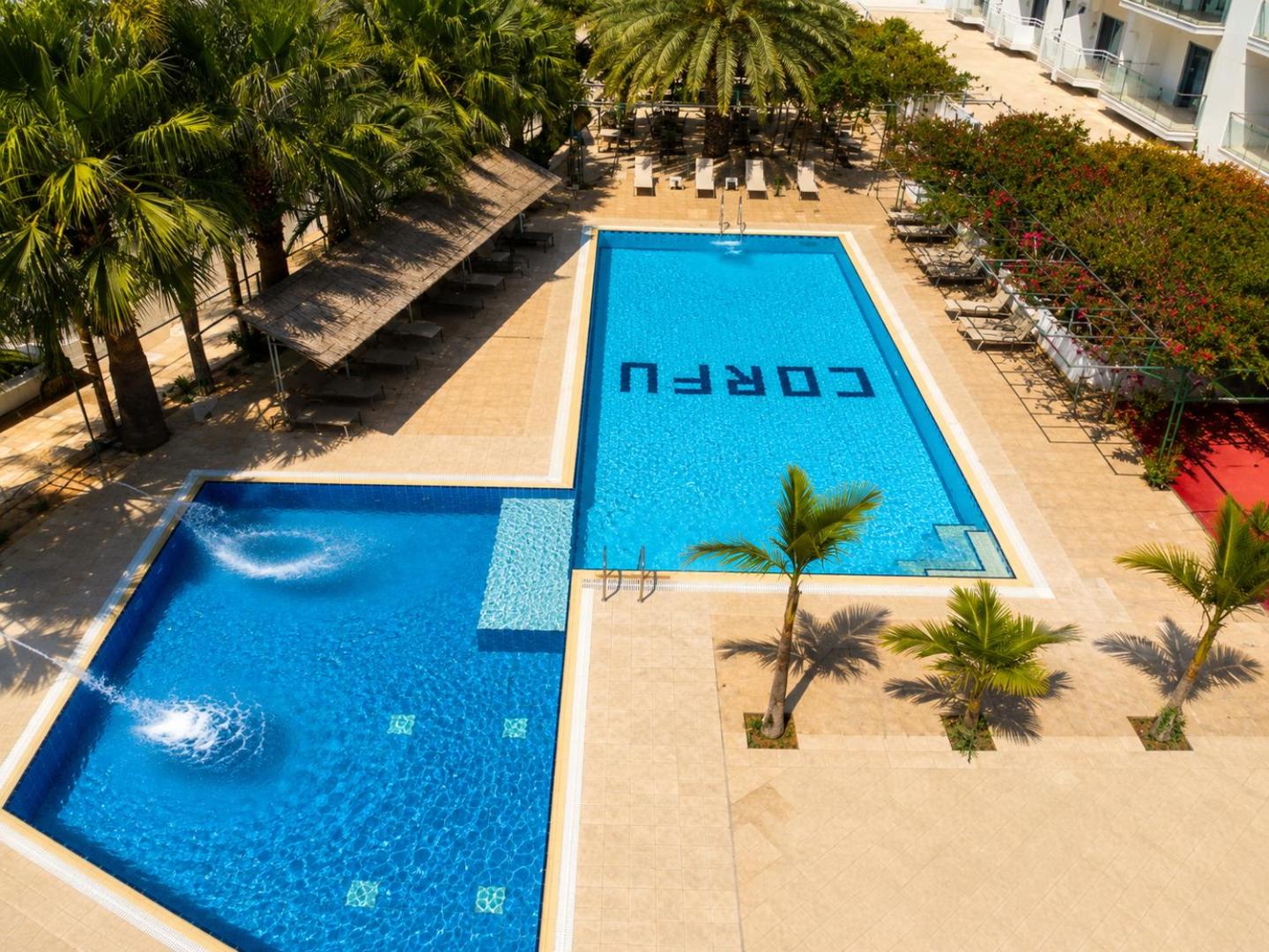 Skats no augšas uz Corfu Hotel baseinu zonu Ajanapā — taisnstūra peldbaseins un bērnu baseins ar strūklakām palmu ielenkumā