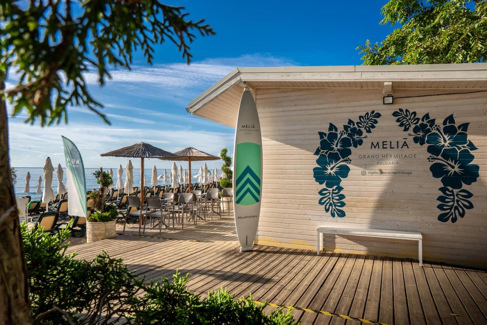 Melia Grand Hermitage pludmales bārs ar koka terasi un sērfošanas dēļa dekoru — atpūtas zona ar saulessargiem pie pašas jūras