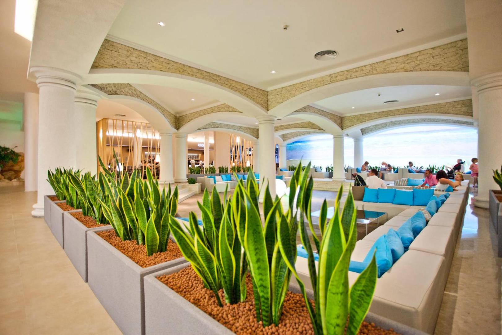 DIT Evrika Beach Club Hotel lobby atpūtas zona ar gaiši ziliem dīvāniem, dekoratīviem augiem podos un arkveida griestu elementiem
