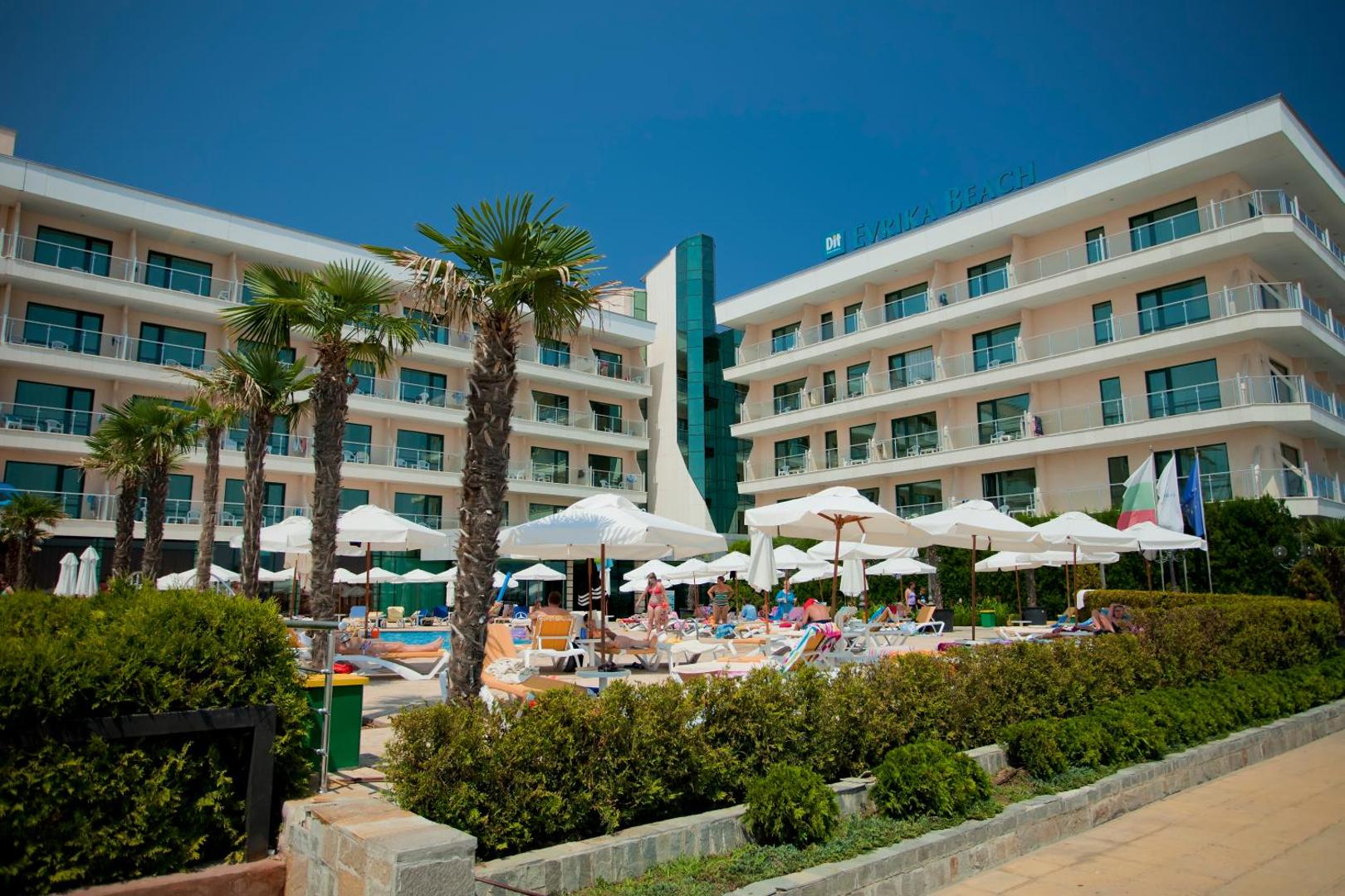 DIT Evrika Beach Club Hotel sauļošanās zona ar baltiem saulessargiem, palmām un atpūtas krēsliem pie baseina