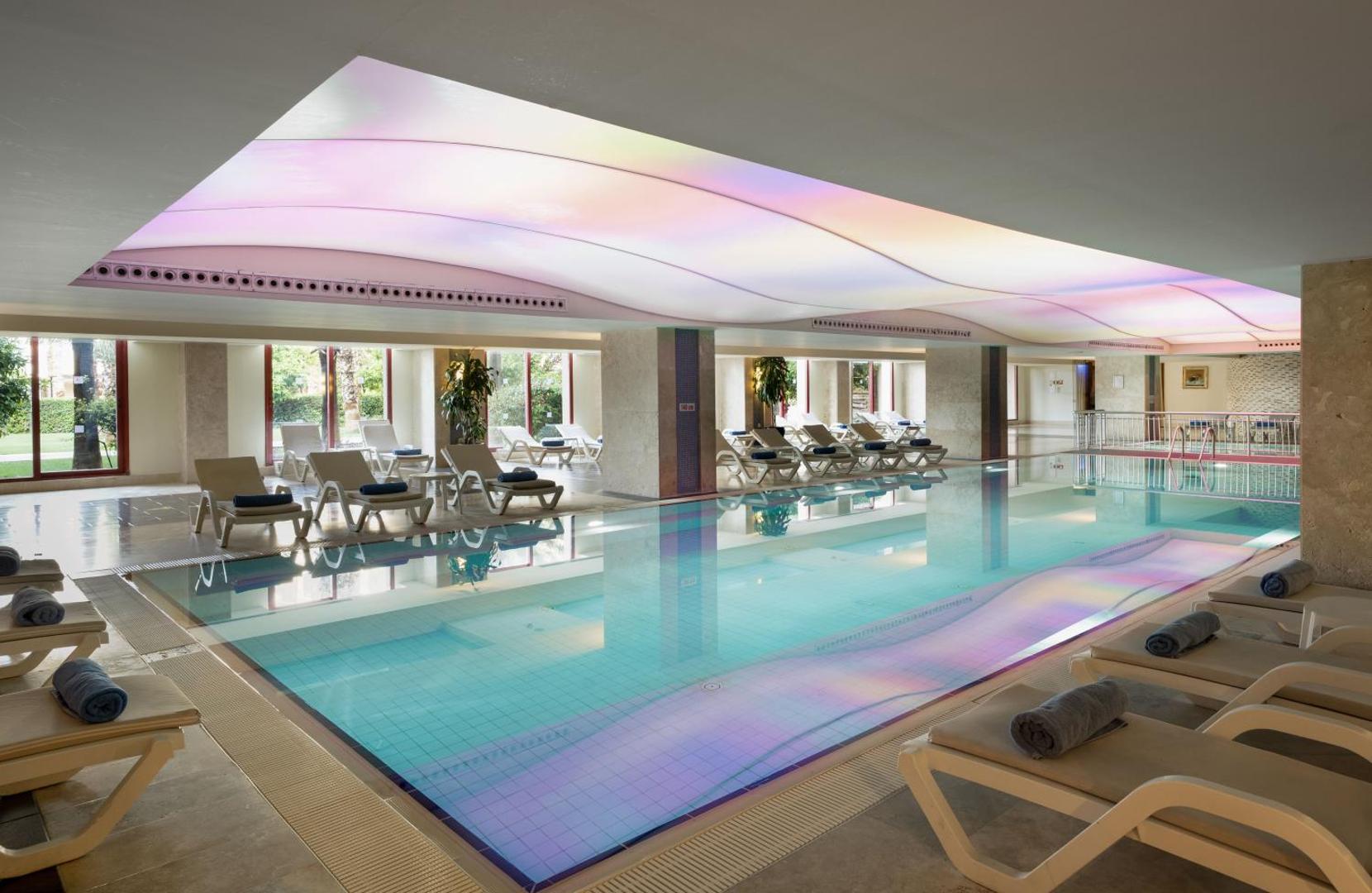 Kaya Belek iekštelpu baseins ar apsildi, atpūtas krēsliem un modernu griestu apgaismojumu viesnīcas spa un fitnesa zonā