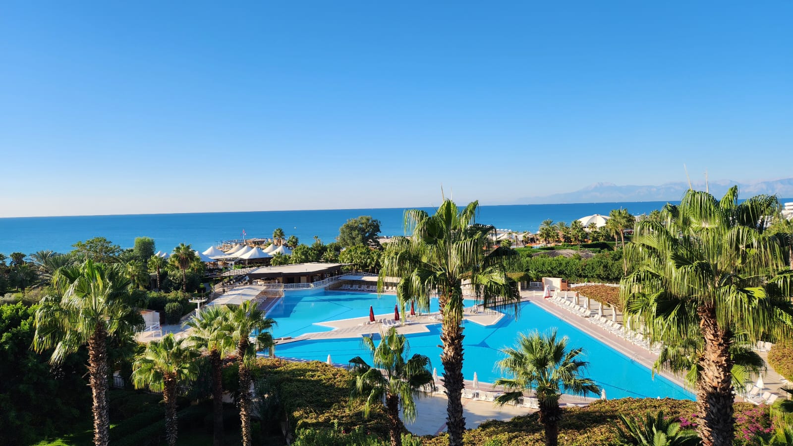 Skats no augšas uz Kaya Belek baseinu kompleksu un palmām Vidusjūras piekrastē, skaidras debesis un kalni tālumā