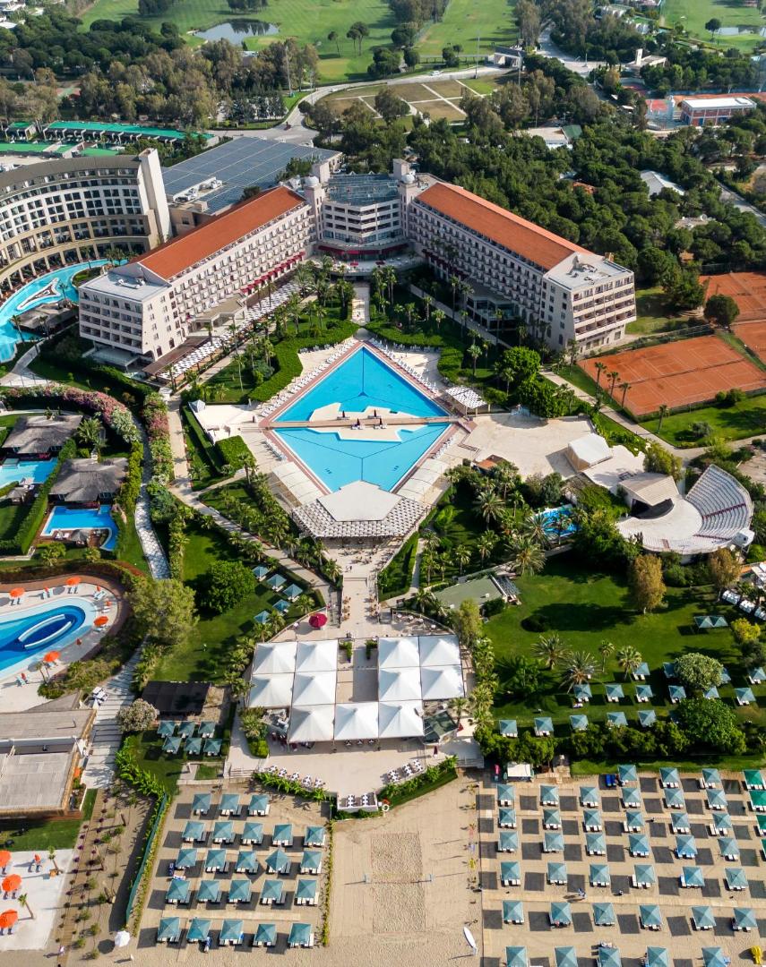 Vertikāls skats uz Kaya Belek centrālo baseinu un viesnīcas korpusiem, ko ieskauj palmas un sakopta dārza ainava