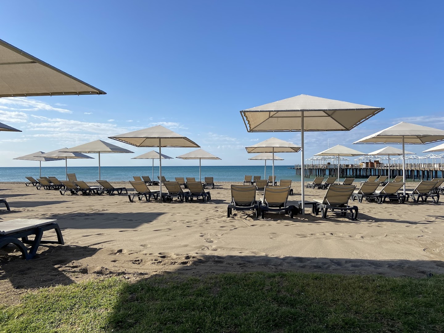 Kaya Belek privātā smilšu pludmale ar gaišiem saulessargiem un ērtiem sauļošanās krēsliem, fonā redzams koka mols un zilās debesis