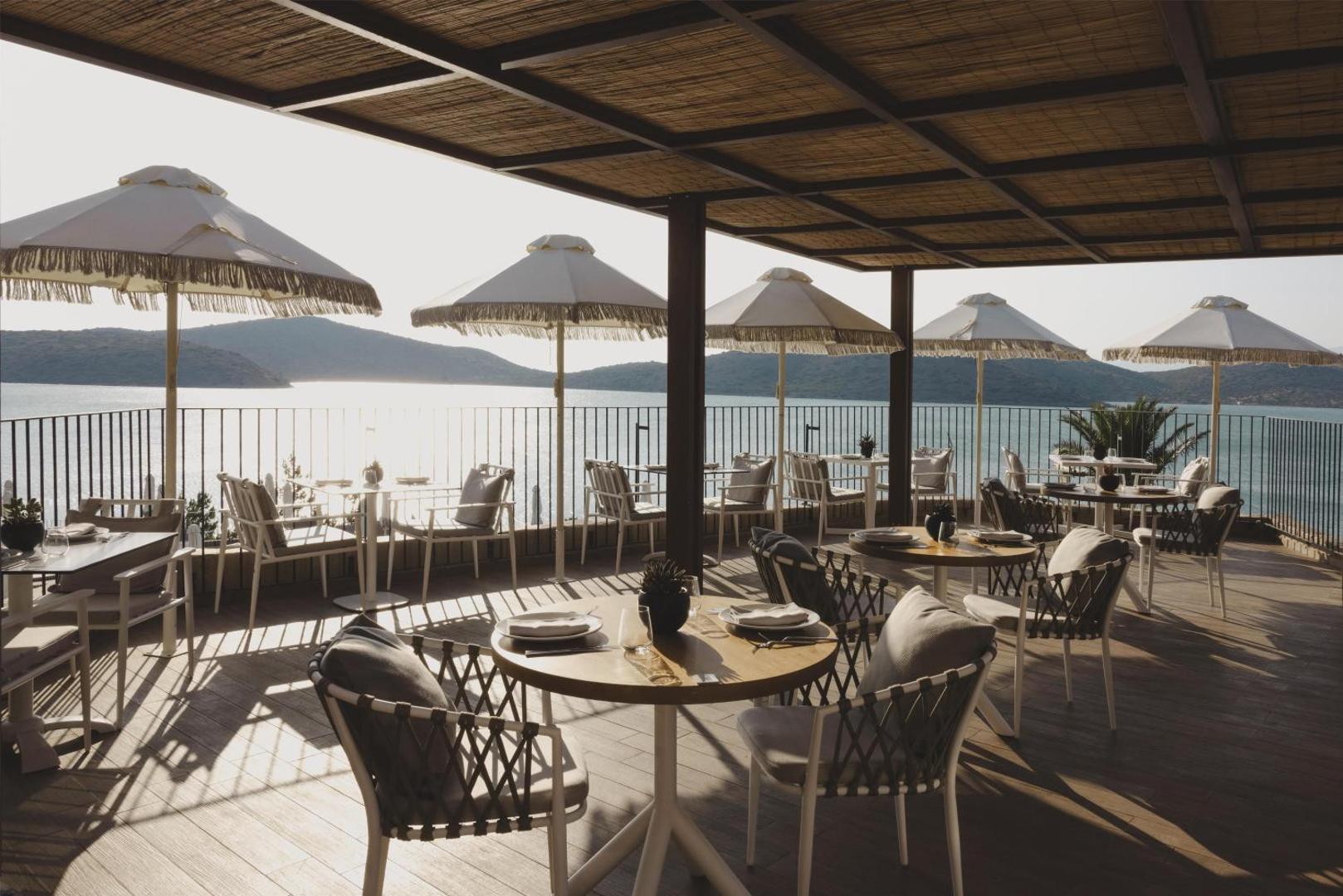 Domes Aulus Elounda Resort āra terases restorāns ar skatu uz jūru un kalniem Krētā — galdiņi zem nojumes ar saulessargiem