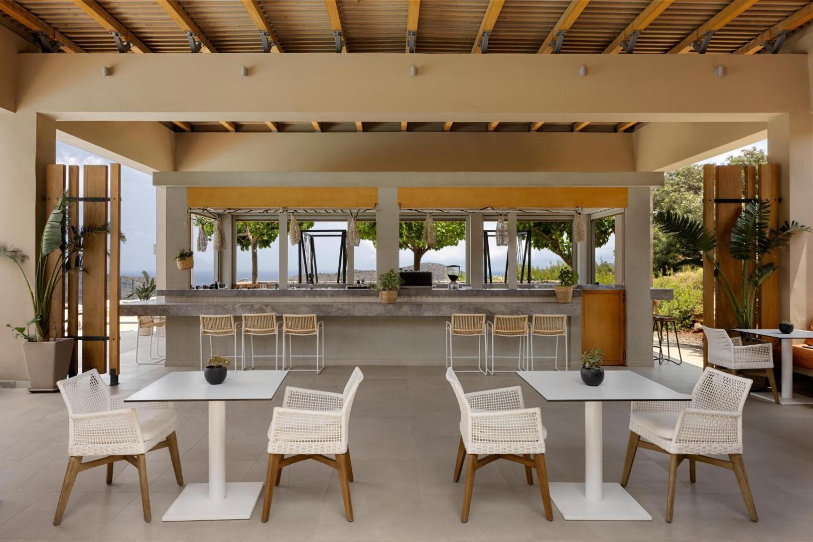 Domes Aulus Elounda Resort bārs ar gaišiem pītiem krēsliem un skatu uz piekrastes dabu Krētas salā