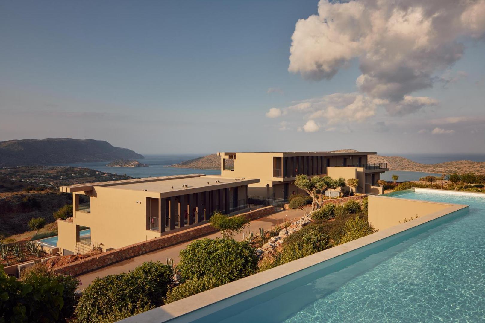 Domes Aulus Elounda Resort baseins ar pārplūdes malu un skatu uz jūru un kalniem — moderna arhitektūra un terases