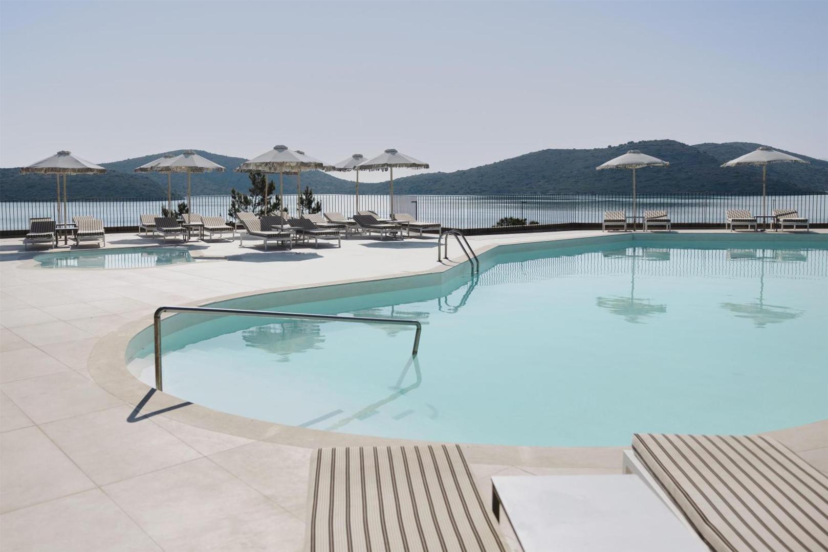 Domes Aulus Elounda Resort baseins ar gaišām flīzēm, saulessargiem un svītrainiem sauļošanās krēsliem piekrastē