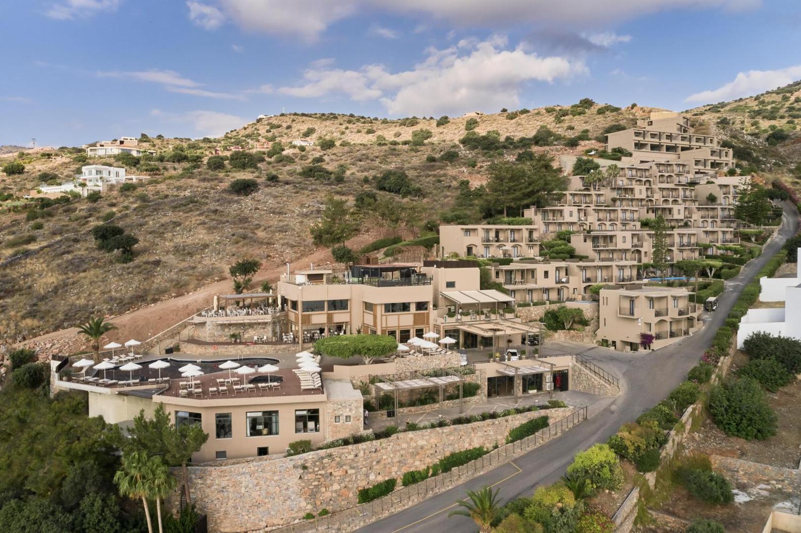 Domes Aulus Elounda Resort galvenā ēka un terases kalna nogāzē — restorānu zonas ar saulessargiem un skatu uz līci