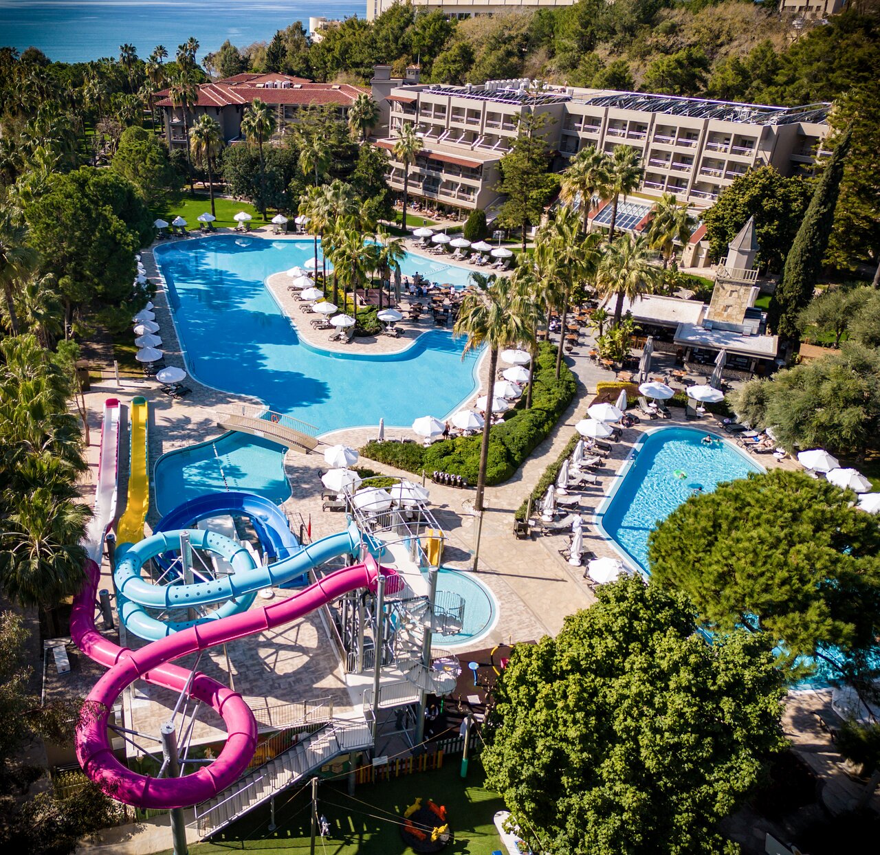 Barut Hemera viesnīcas teritorijas skats no augšas ar vairākiem baseiniem, krāsainiem ūdens slidkalniņiem un zaļojošu dārzu.