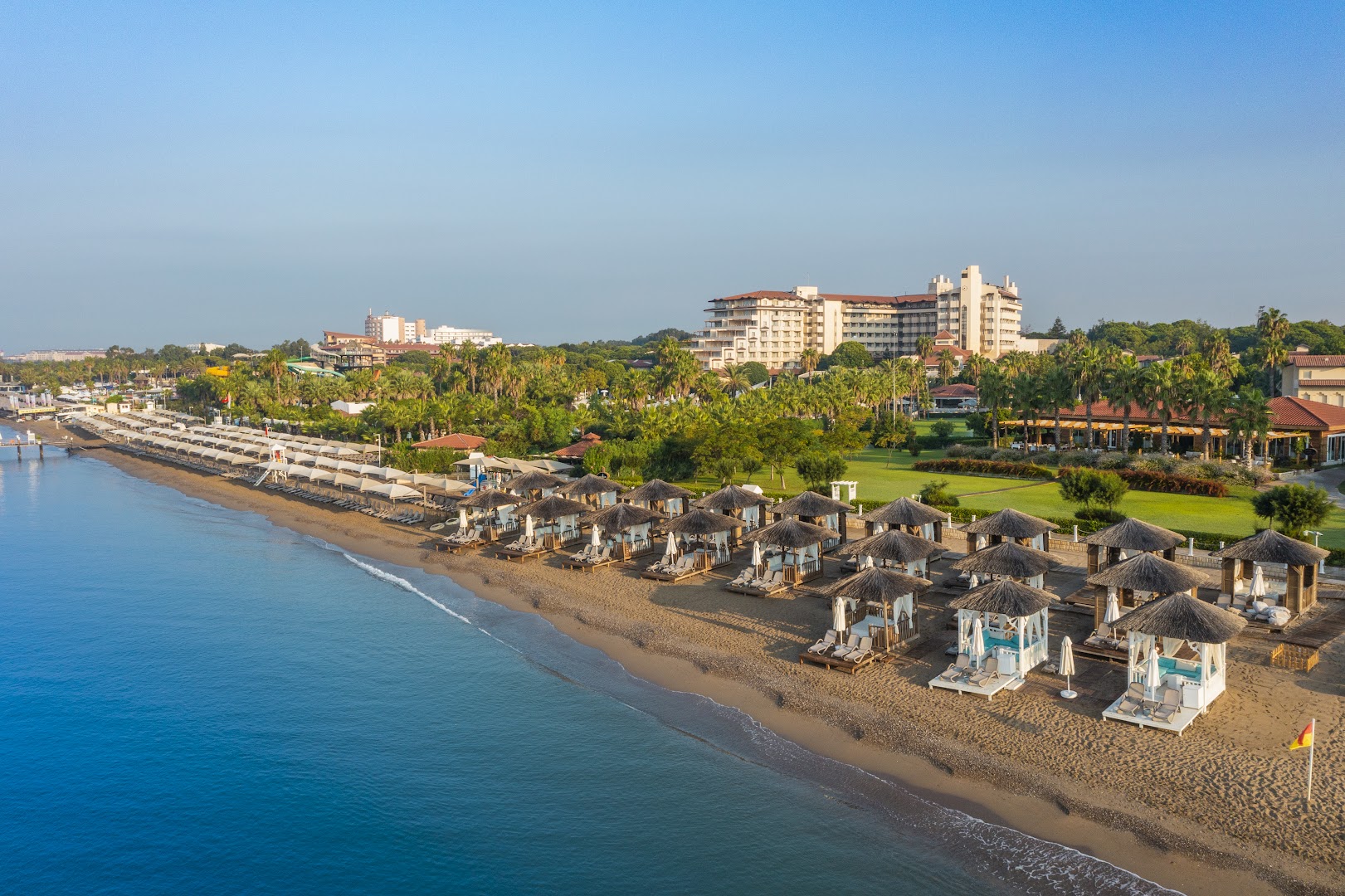 Bellis Deluxe Hotel panorāmas skats uz privāto pludmali Belekā — tirkīza jūra, smilšu krasts ar saulessargiem un viesnīcas ēka fonā