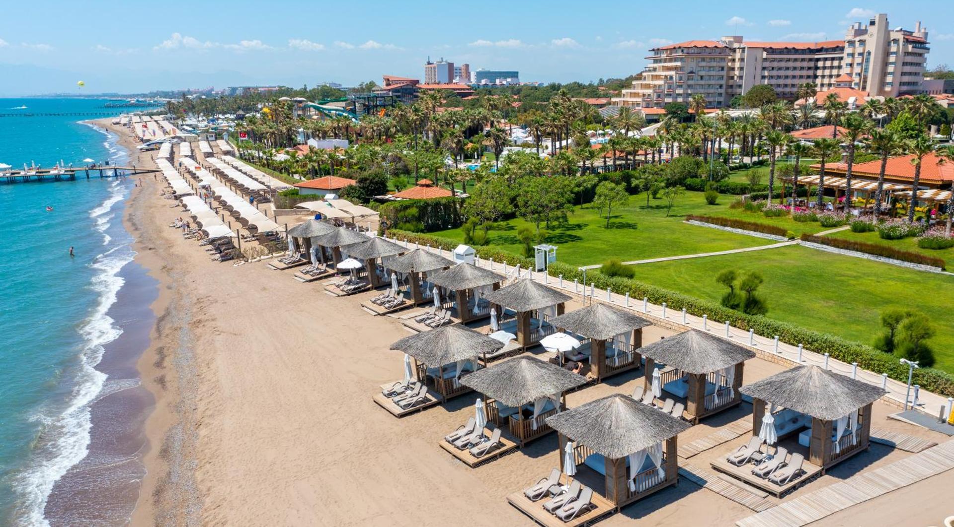 Bellis Deluxe Hotel pludmales zona no augšas — sakopts zāliens, palmas, smilšu josla un rinda ar komfortablām atpūtas nojumēm