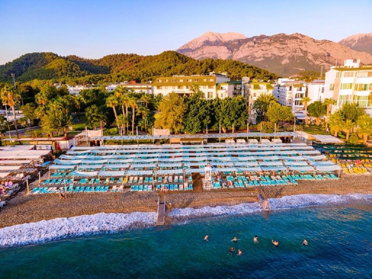 L'ancora Beach Hotel skats no putna lidojuma uz oļu pludmali, ziliem saulessargiem un kalnu fonu Kemerā, Turcijā