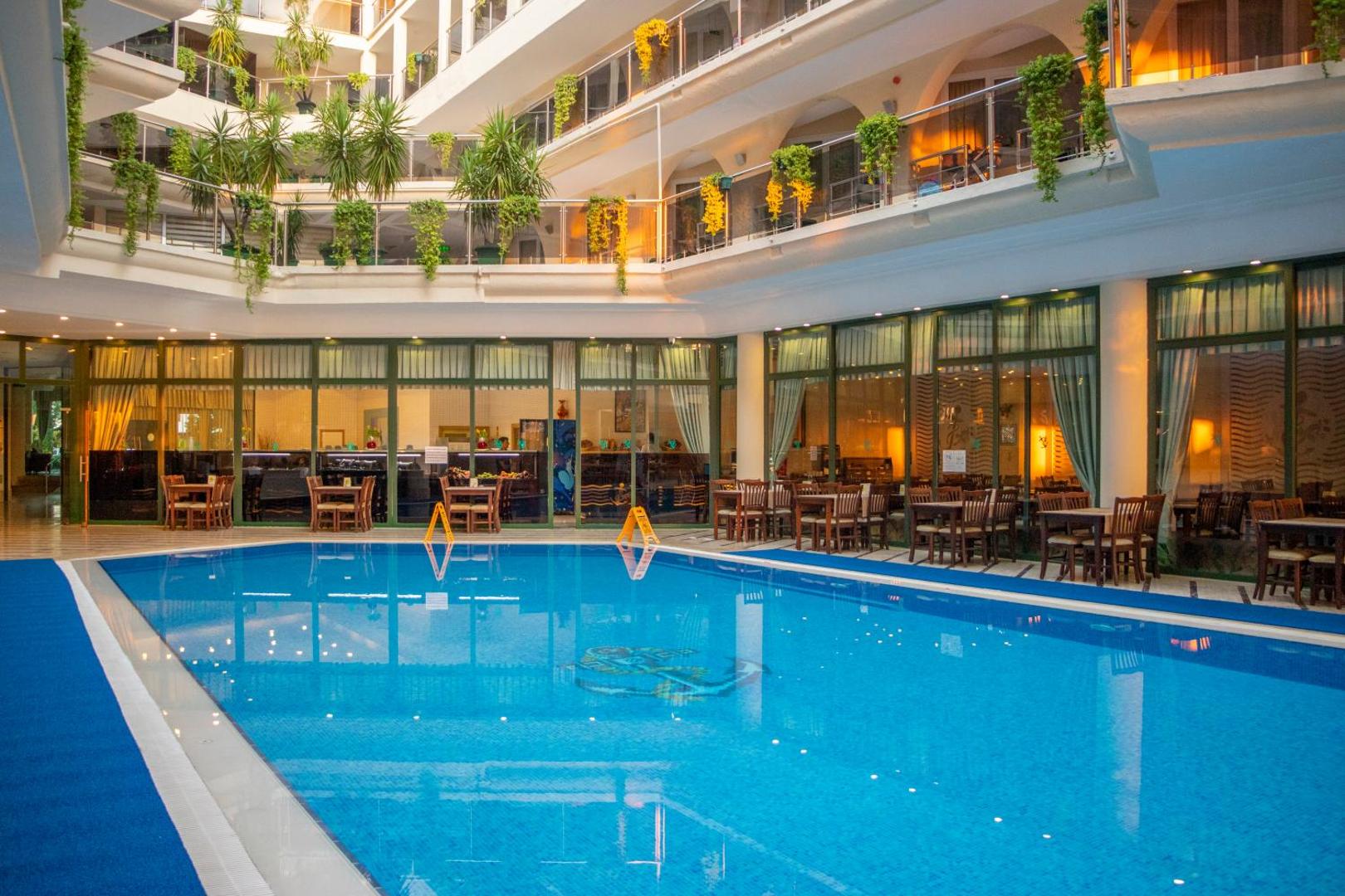 L'ancora Beach Hotel iekšpagalma baseins vakara apgaismojumā — apkārt restorāna galdiņi un viesnīcas korpusi ar apzaļumotiem balkoniem