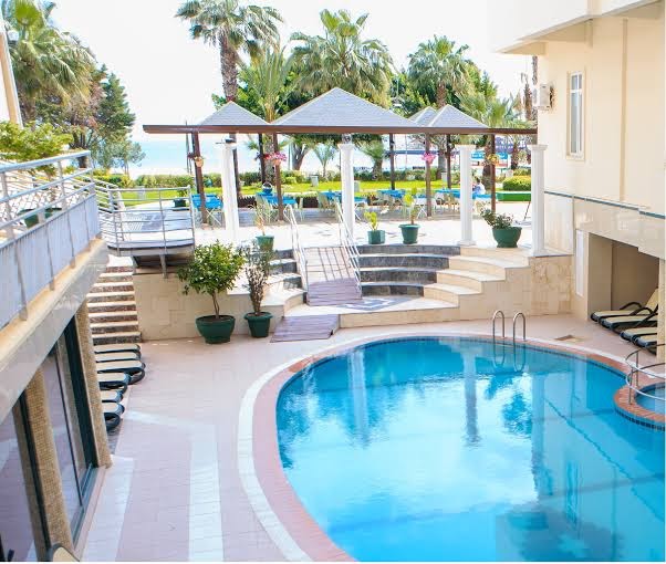 L'ancora Beach Hotel āra baseins ar saulessargiem un palmām piekrastē — fonā redzama Vidusjūra un kalnu ainava