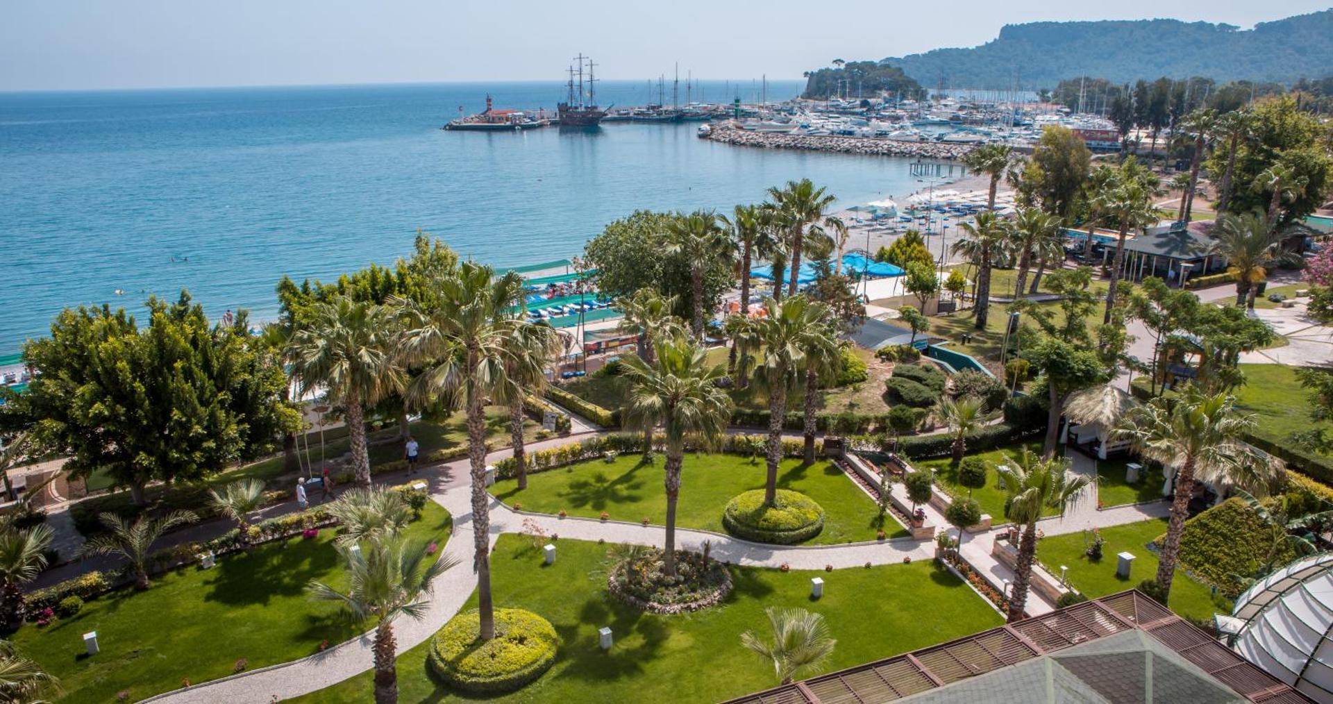 Panorāmas skats no L'ancora Beach Hotel uz Kemeras ostu un Vidusjūru — kopti dārzi ar palmām un jahtu piestātne fonā