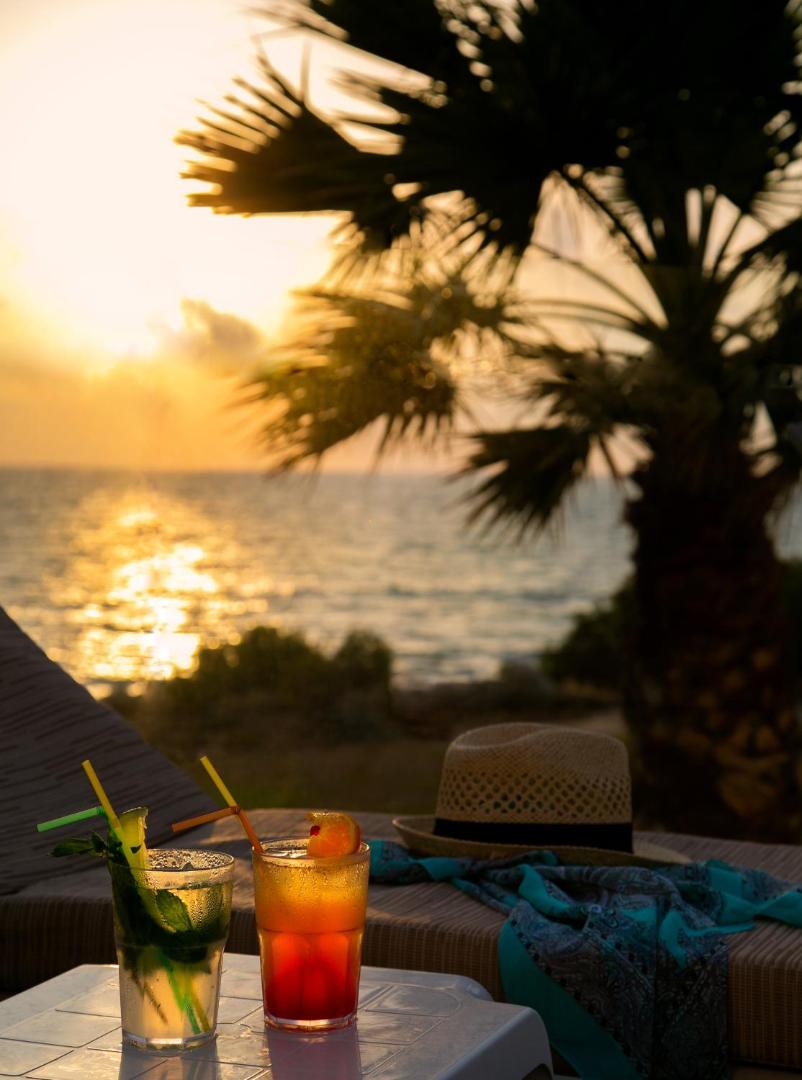 Saulrieta skats no Louis Paphos Breeze atpūtas zonas Pafosā — kokteiļi uz galdiņa, palmas un Vidusjūras panorāma vakarā