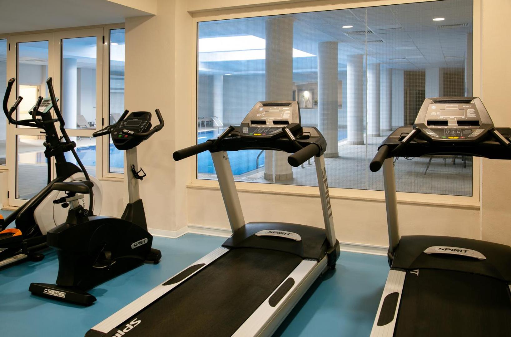 Louis Paphos Breeze fitnesa centrs ar moderniem skrejceliņiem un velotrenažieriem, fonā redzams iekštelpu baseins caur lieliem logiem