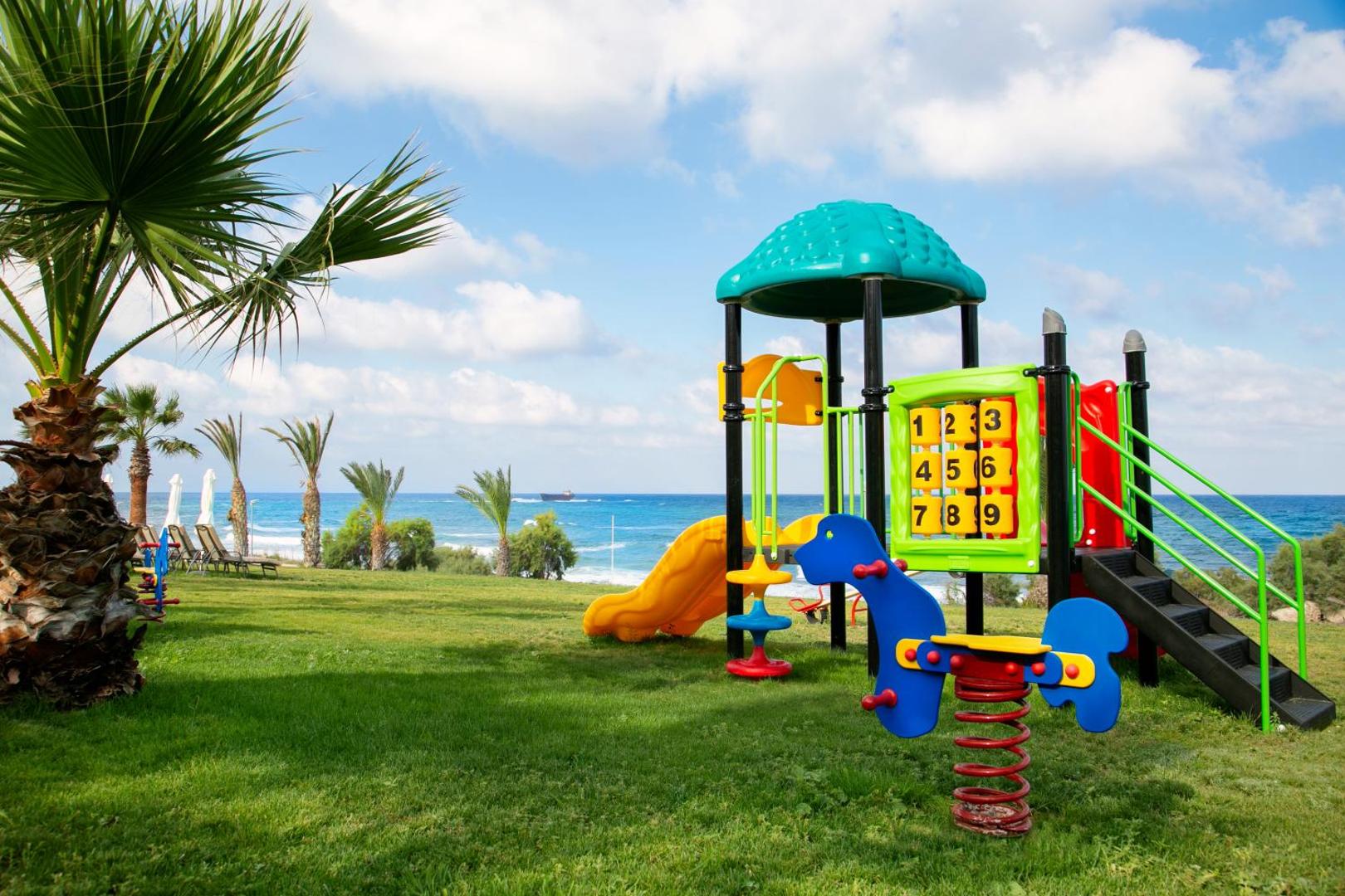 Bērnu izklaides zona Louis Paphos Breeze viesnīcā Pafosā, Kiprā — krāsains rotaļu namiņš un slidkalniņš uz zaļa zālāja