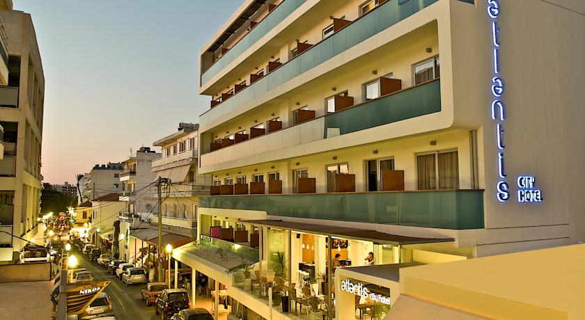 Plašs skats uz Atlantis Boutique City Hotel fasādi un Rodas pilsētas ielu saulrietā — viesnīcas terase un apkārtējā pilsētvide.
