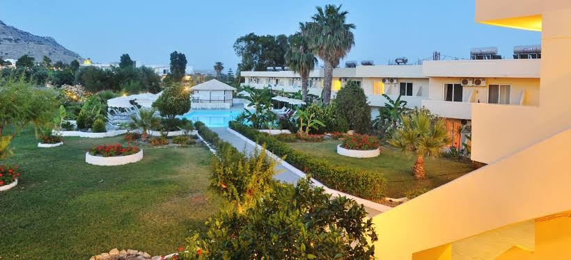 Olive Garden Hotel Rodā skats no balkona uz dārzu, palmām un baseinu tālumā — zaļš mauriņš un viesnīcas arhitektūra