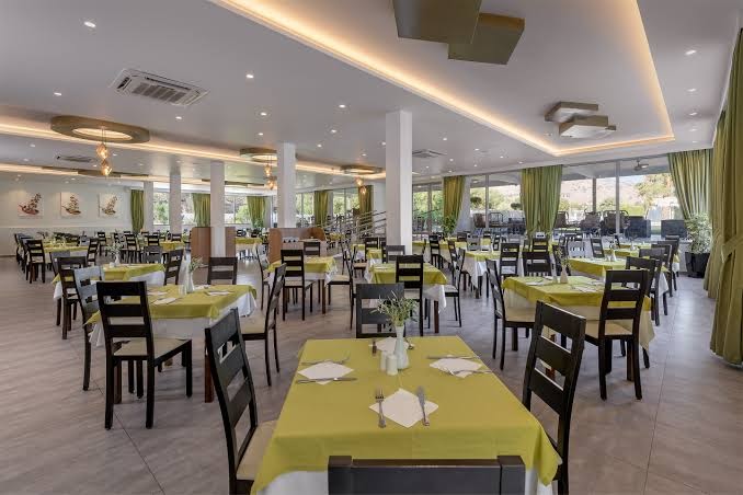Gaiša un plaša Olive Garden Hotel restorāna zāle ar klātiem galdiem, zaļiem akcentiem un skatu uz dārzu