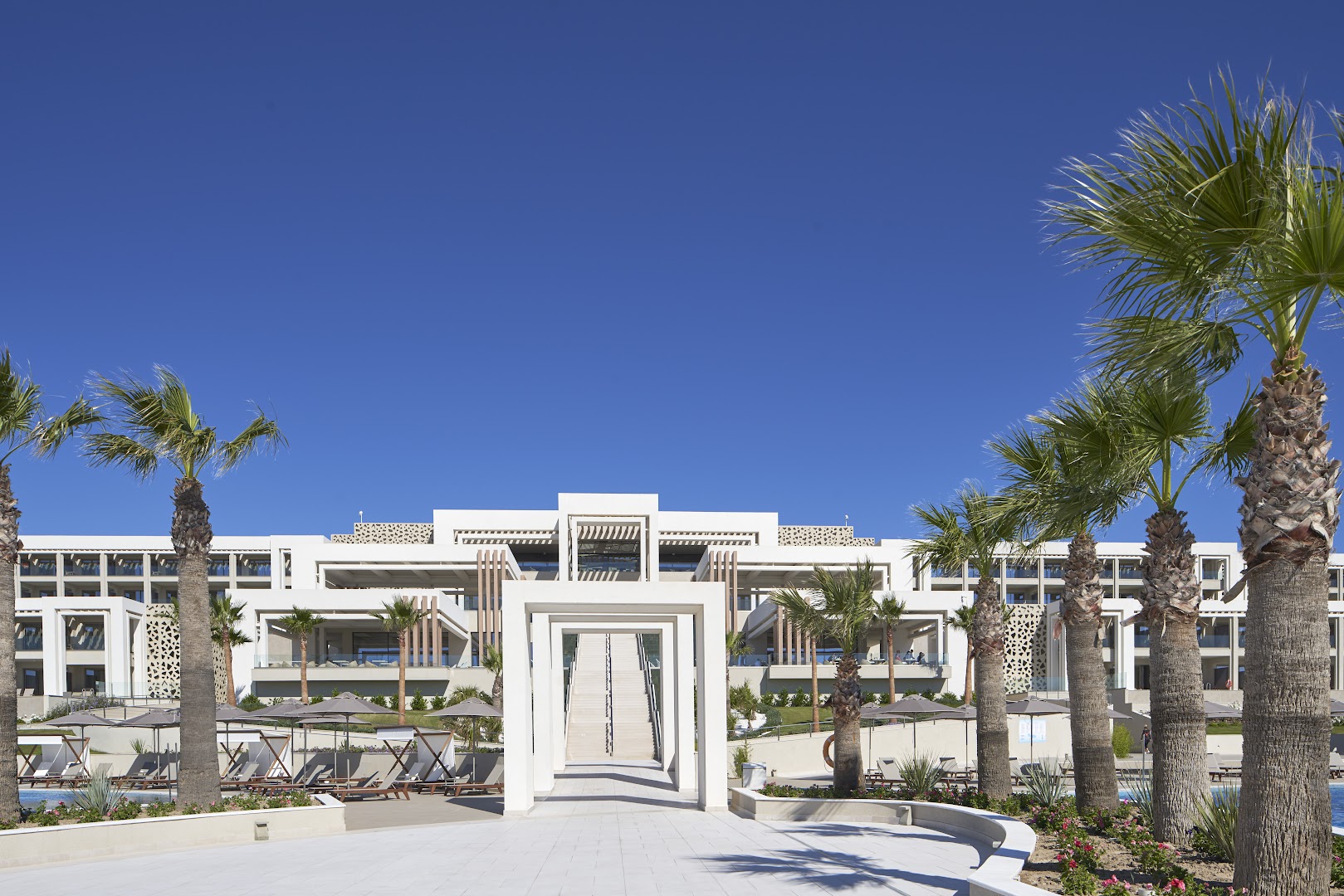 Mayia Exclusive Resort & SPA centrālā eja ar palmām un modernām baltām arkām uz galvenās ēkas fonu skaidrās debesīs